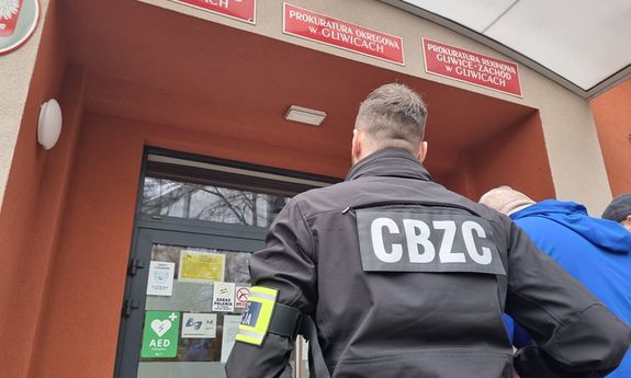 policjant ma na kurtce napis CBZC. Prowadzi do budynku zatrzymanego mężczyznę