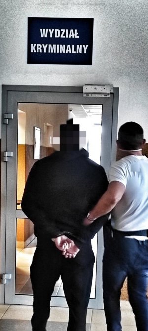 Mężczyzna - policjant bez munduru prowadzi mężczyznę w mundurze, w tle korytarz i szyld Wydział Kryminalny
