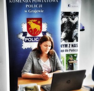 Kobieta rozmawiająca online z uczniami, w tle baner z napisem Komenda Powiatowa Policji w Grajewie i logo powiatu grajewskiego w postaci łosia.