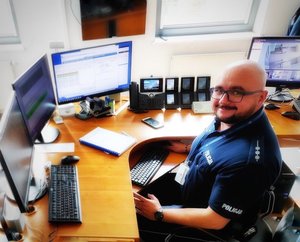 Policjant dyżurny na stanowisku kierowania jednostką, przed nim monitory komputerów.