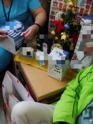 Pielęgniarka, policjant bez munduru przekazujący pielęgniarce produkty medyczne na przykład inhalator, urządzenie do mierzenia pulsu. W tle choinka, kolorowe torby świąteczne na prezenty.
