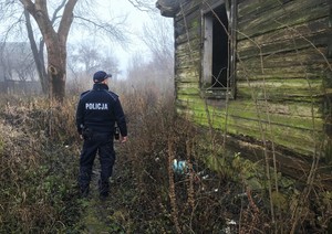 Policjant stojący tyłem, patrzy w stronę drewnianego budynku